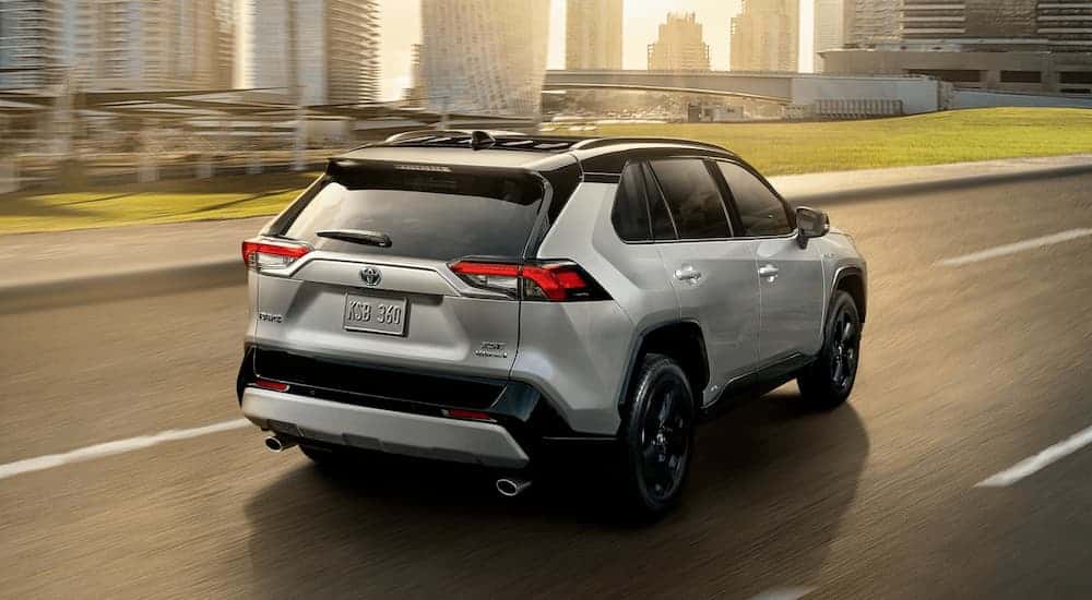 A grey 2020 Toyota RAV4 is driving toward a city with the sun shining through buildings.