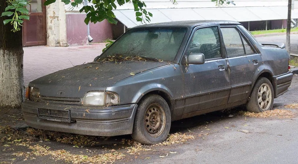 An old, beat up, gray car is parked on a street covered with tree debris.