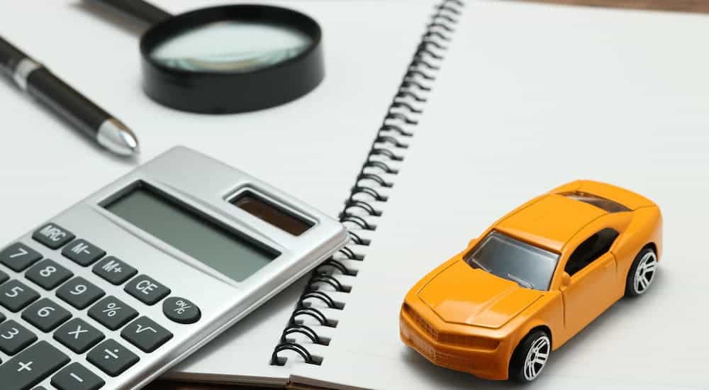 An orange toy car is sitting on a notebook with a calculator and magnifying glass.