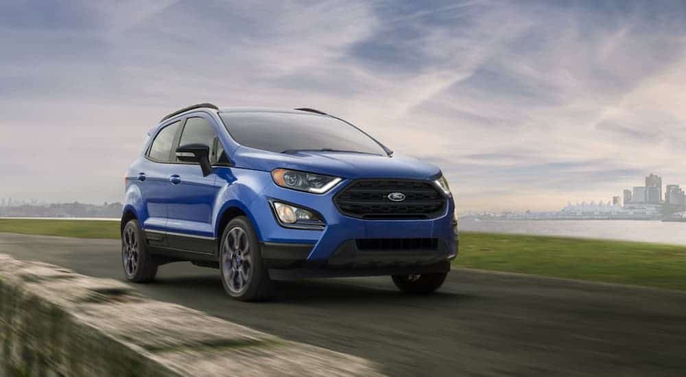 A blue 2020 Ford EcoSport is driving past a stone wall with a city skyline in the distance.