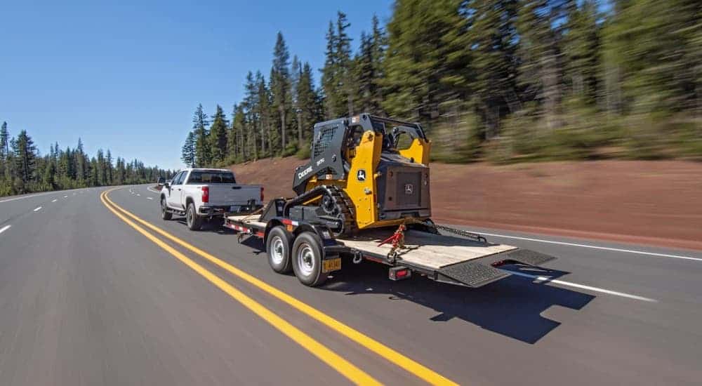 A white 2020 Chevy Silverado 2500HD is towing a trailer with a skid-steer on a tree-lined road.
