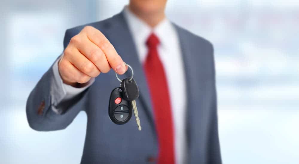 A car salesman is holding out a car key to the viewer.