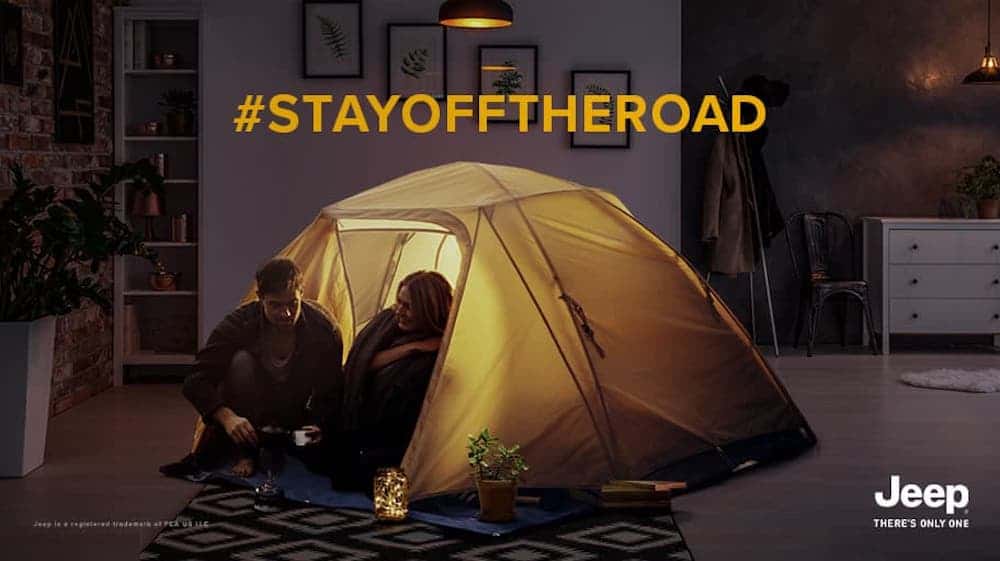 A man and woman are camping in a tent inside a living room with the text "#stayofftheroad".