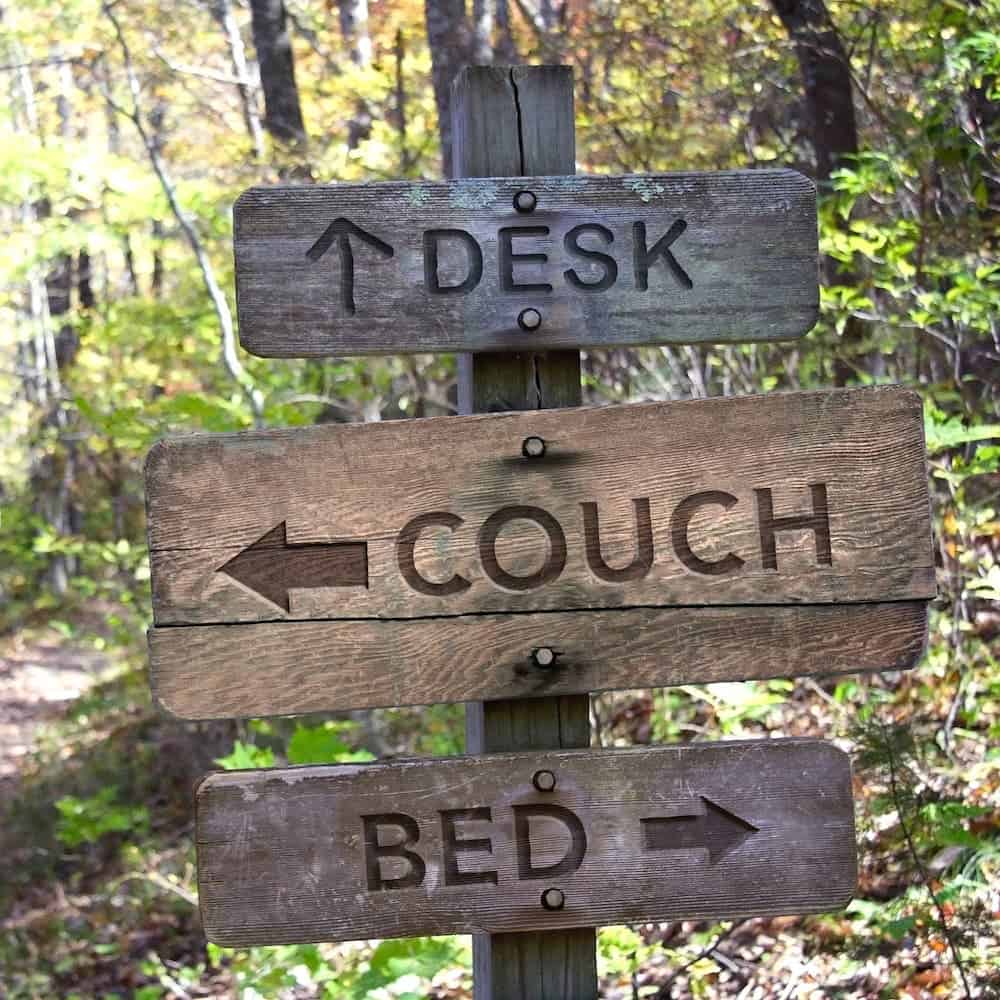 Three wooden signs are on a trail in the woods and read "Desk, Couch, Bed" with arrows pointing in different directions.