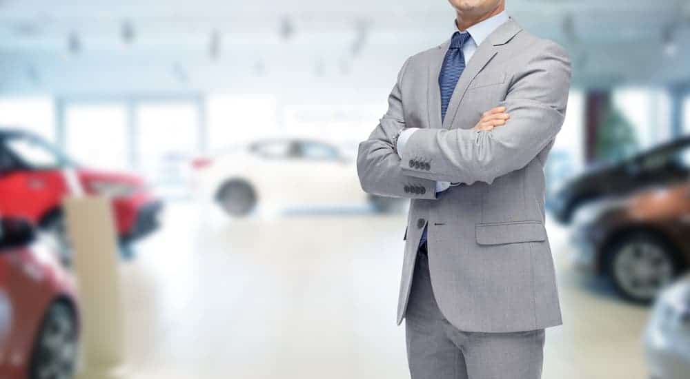 A car salesman in a grey suit has his arms crossed.