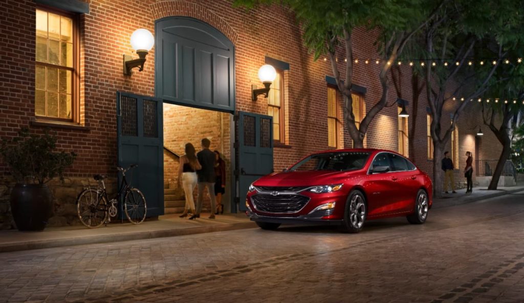 A red 2020 Chevy Malibu is parked in front of a red brick building at night after winning the 2020 Chevy Malibu vs 2020 Nissan Altima comparison.