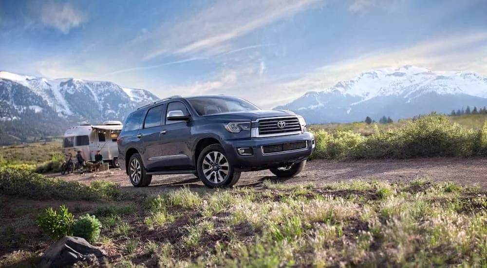 A blue 2020 Toyota Sequoia is parked in front of a campsite and silver Airstream camper with mountains in the distance.