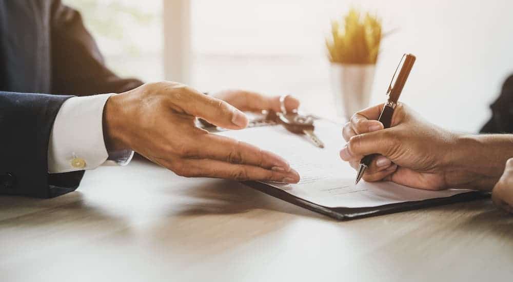 A closeup shows two pairs of hands signing an auto loan paperwork next to car keys.