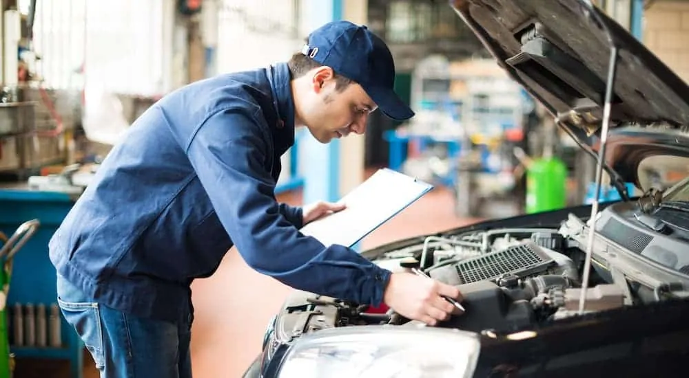 A mechanic is leaning over an engine bay and holding a clipboard.