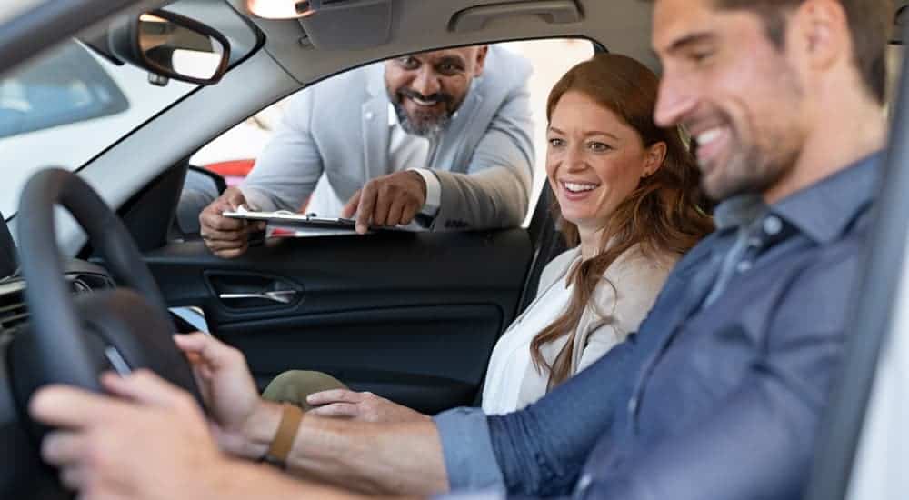 A salesman is talking to a couple in a car at a local used car dealership.