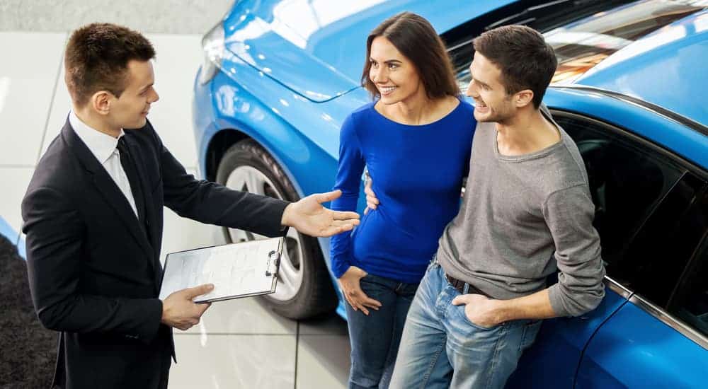 A salesman is talking to a couple while they lean on a blue car.