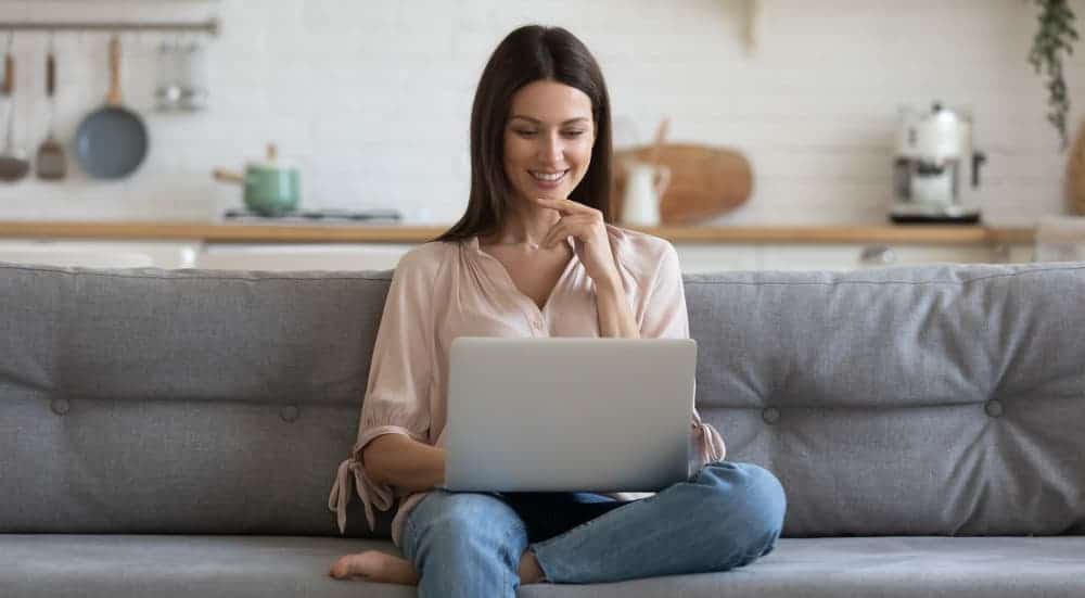 A woman is sitting on the couch and smiling while on her laptop.