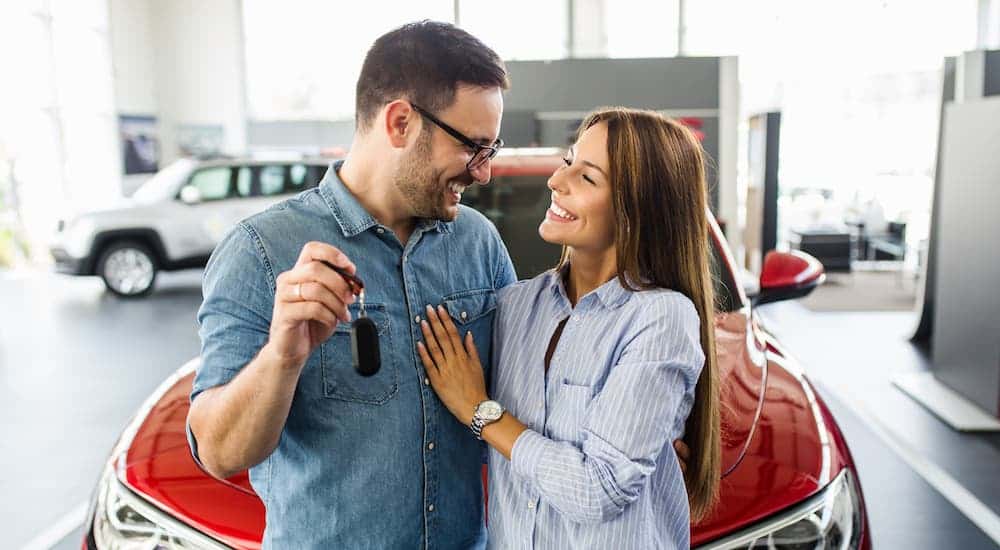 A happy couple is sitting on the hood of a car and holding keys.