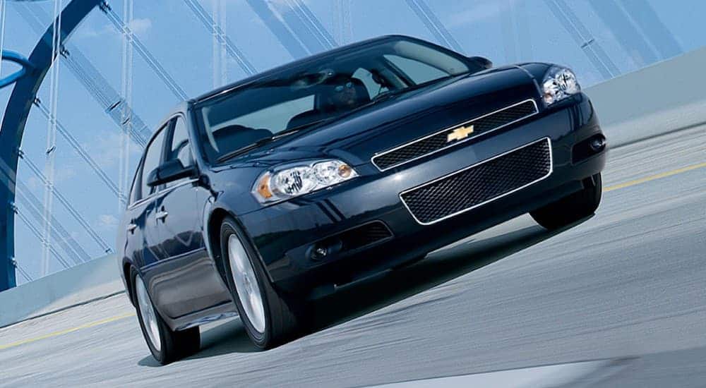 A dark grey 2012 Chevy Impala is driving over a bridge, shown from a slanted angle.