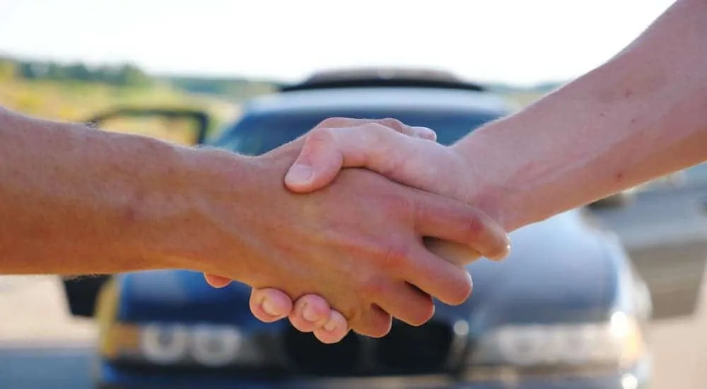 A close up is shown of two hands shaking in front of a parked car.