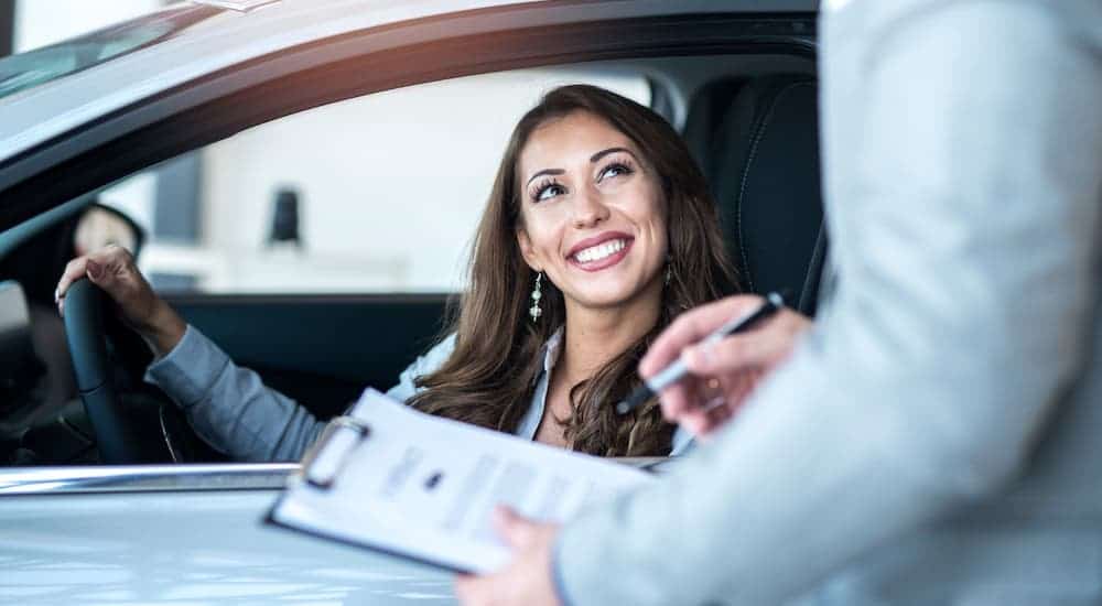 A woman is sitting in a car for sale near you and talking to a salesman holding a clipboard.