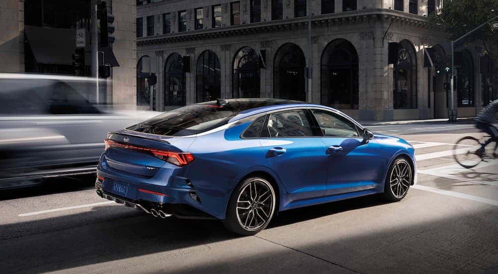 A blue 2021 Kia K5 is stopped at an intersection on a busy city street.