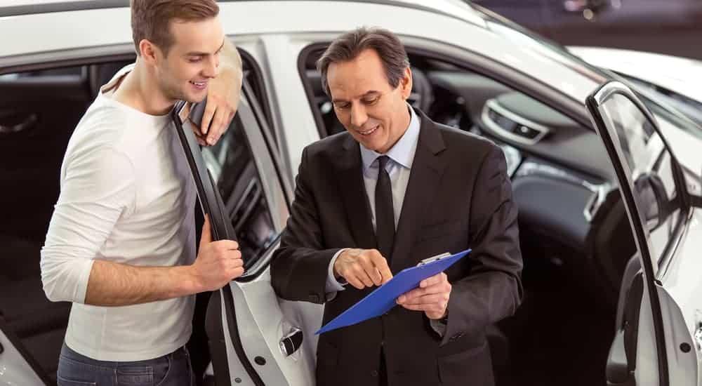 A salesman is showing a man a clipboard with information on affordable used cars while they stand in front of a white vehicle.