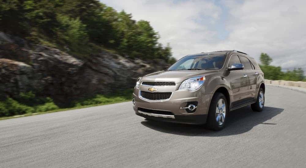 A beige 2014 Chevy Equinox is driving past a rocky hill and trees.