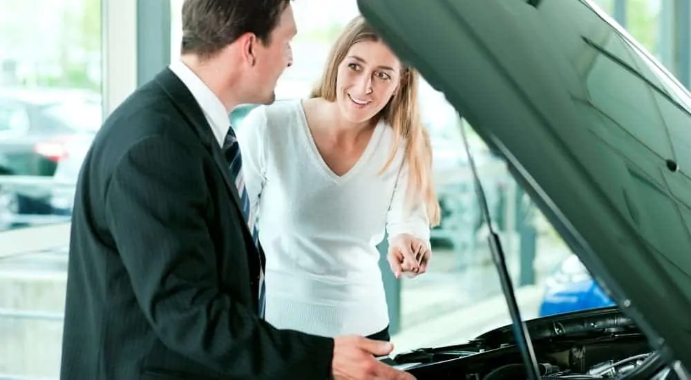 A woman is looking under the hood of a car next to a salesman at a used car lot.