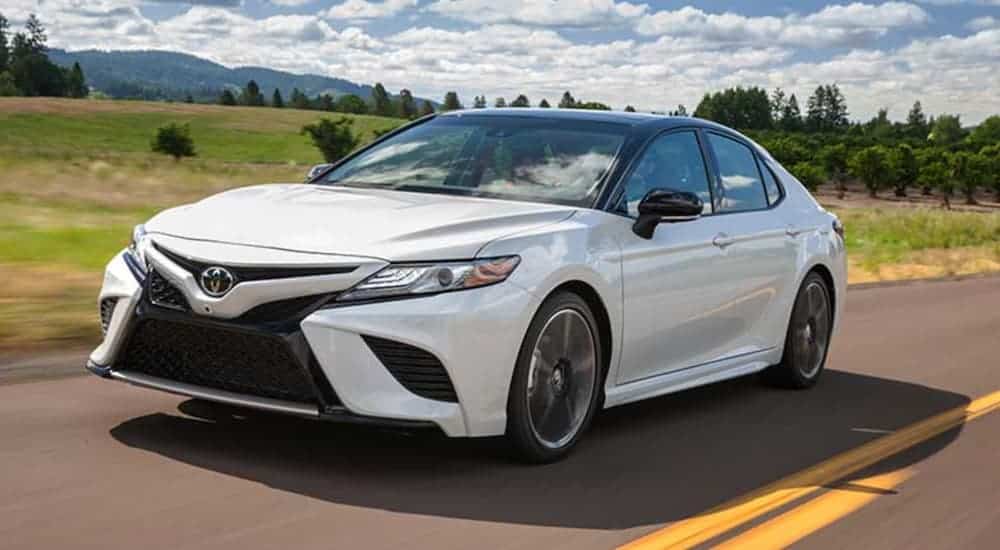 A white 2018 Toyota Camry is driving past a field and distant mountain.