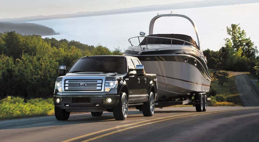 A black 2014 Used Ford F-150 is towing a large boat uphill with a lake in the distance.