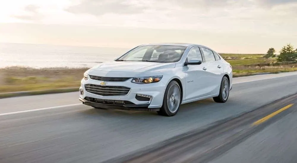 A white 2016 Chevy Malibu is driving past the ocean on an overcast day.