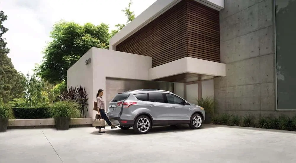 A silver 2013 used Ford Escape is parked in front of a modern house with a woman opening the lift gate with a foot sensor.