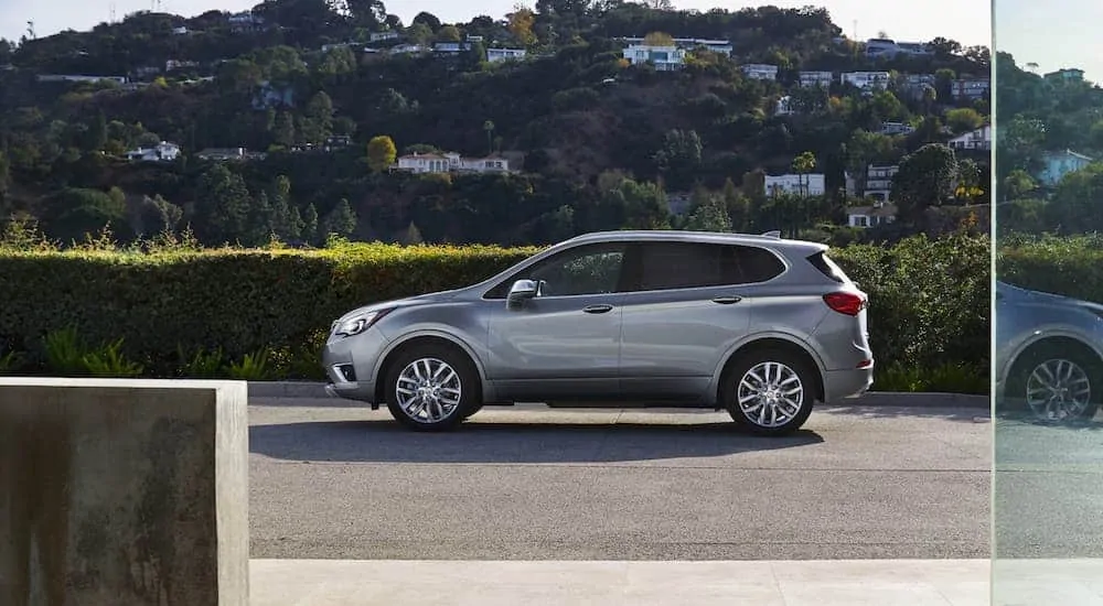 A grey 2020 Buick Envision is parked in front of a hedge with many houses on a hill behind it.