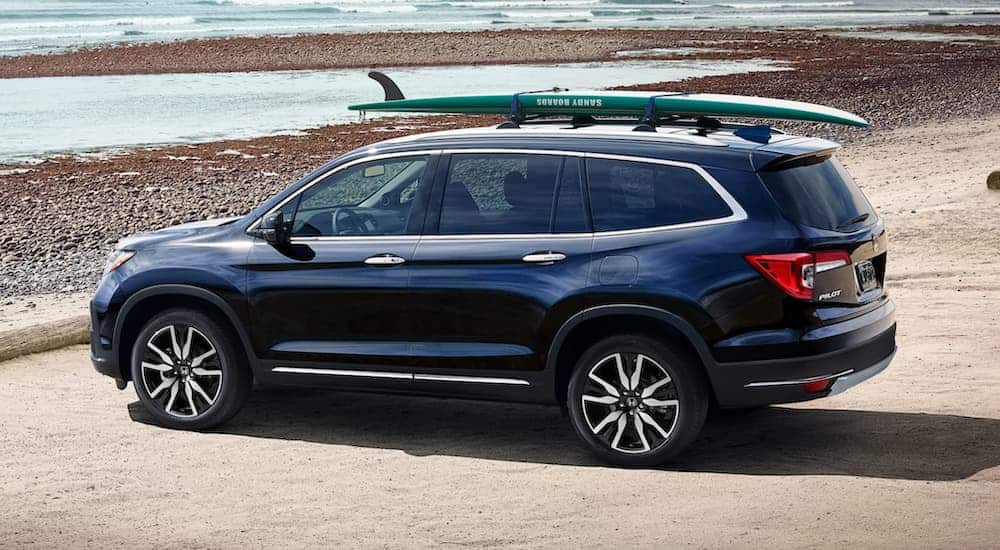 A blue 2021 Honda Pilot is parked next to a rocky beach with a surf board on the roof.