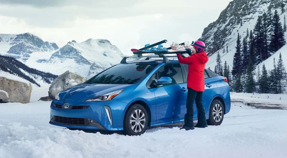 A blue 2021 Toyota Prius XLE is parked on a snowy mountain and woman is loading a snowboard on to the roof rack.