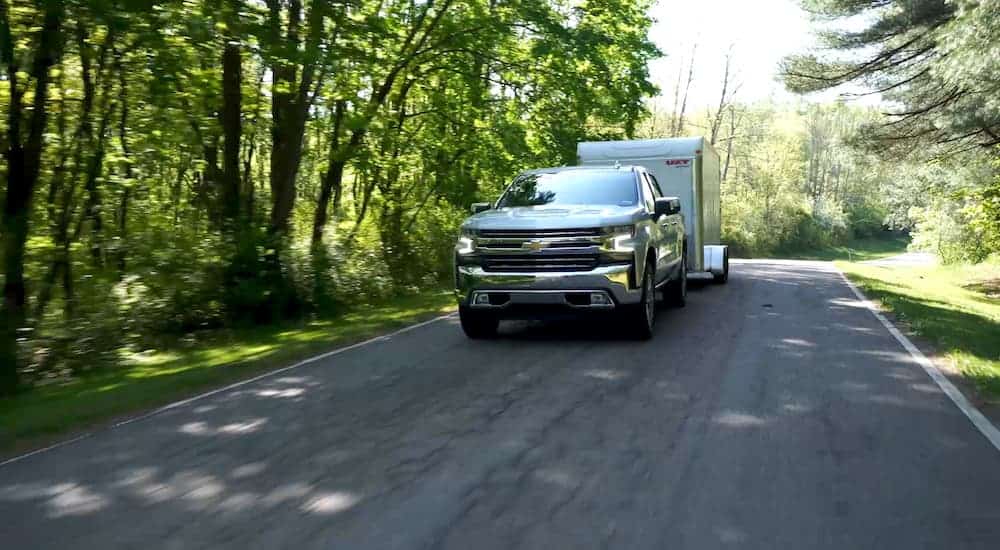 A silver 2019 used Chevy Silverado 1500 is towing a enclosed trailer down a tree lined road.