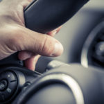 Close up of a hand wrapped around the top left of a black steering wheel