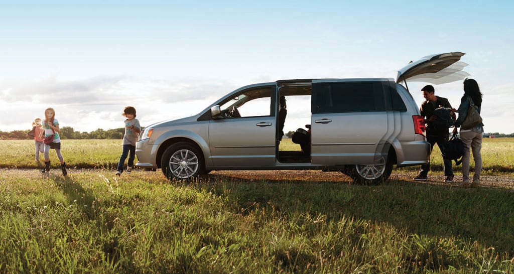 A family loading up a silver Dodge Caravan in a grassy field against a blue sky