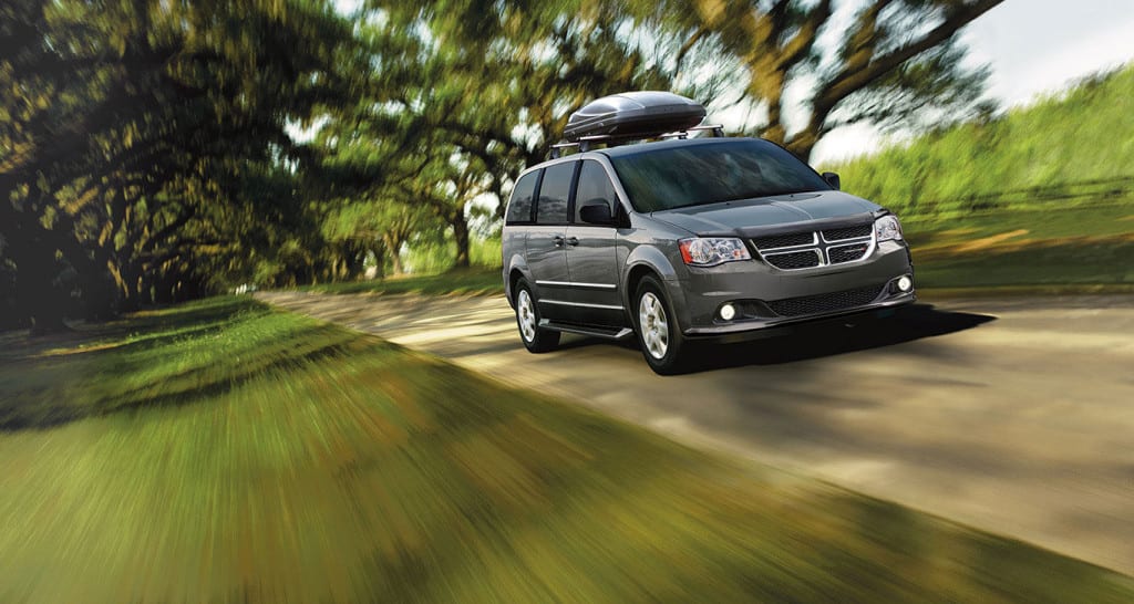 A grey 2015 Dodge Grand Caravan with roof cargo carrier is driving on a tree lined road.