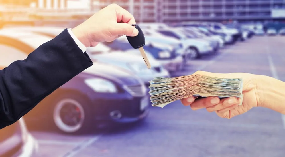 Hands in front of a row of vehicles, one holding a key and the other holding a stack of cash