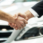 Man in a suit shaking hands with a man in a long sleeve shirt in front of the grill of a white vehicle