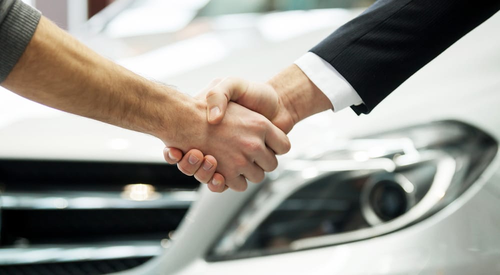 Man in a suit shaking hands with a man in a long sleeve shirt in front of the grill of a white vehicle