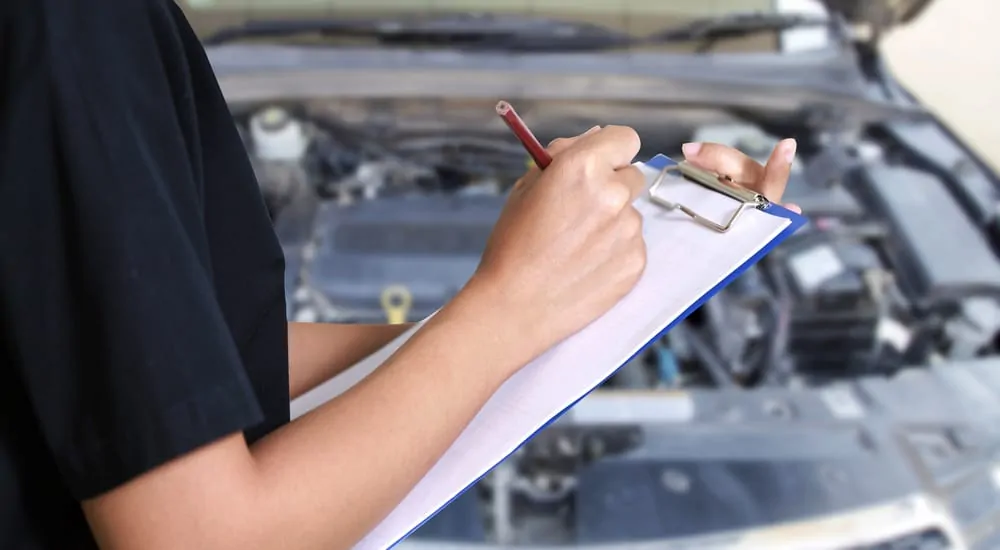 Person in a black shirt holding a clipboard and a red pencil in front of a car with the hood open