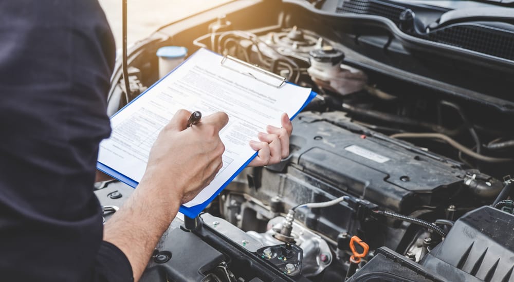 Person in blue shirt holding a clipboard over the open hood of a car