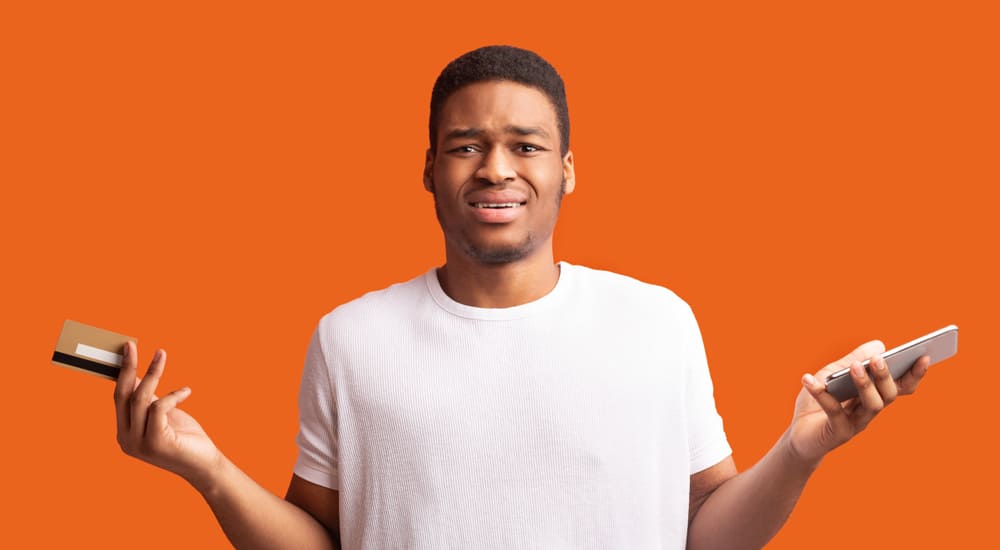 Man in a white shirt against an orange background, shrugging with a credit card in one hand and a smartphone in the other