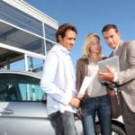 A salesman shows a couple car information on a tablet against a blue sky