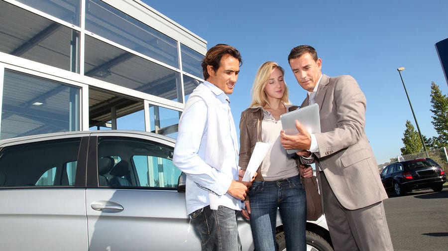 A salesman shows a couple car information on a tablet against a blue sky