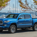 A blue 2018 Toyota Tacoma is parked in front of a bay with sailboats.