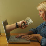 Hand coming out of a silver laptop, handing dollar bills to a blonde haired woman in a blue shirt