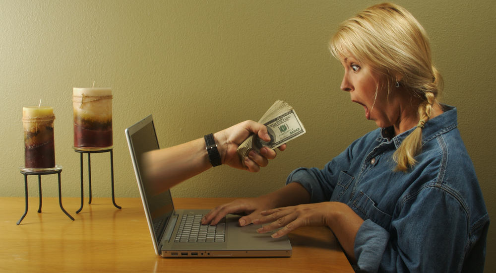 Hand coming out of a silver laptop, handing dollar bills to a blonde haired woman in a blue shirt