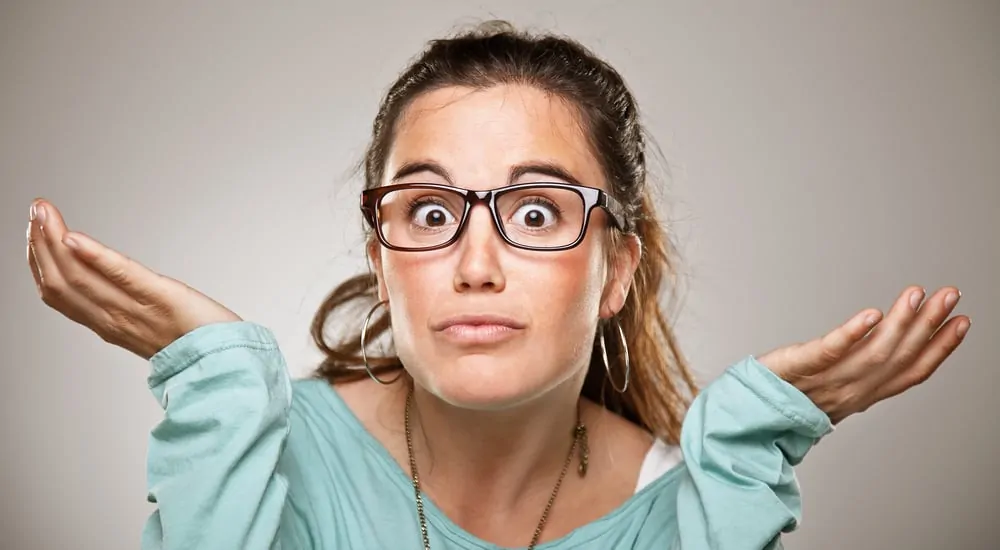 Woman in a light blue shirt and glasses shrugging with her eyes wide open