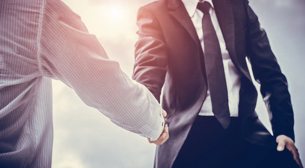 Man in a black suit, white shirt, and tie shaking hands with a man in a button-up