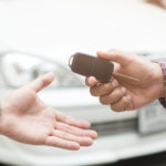 Man's arm wearing a plaid shirt handing a car key to another man's hand in front of a white car