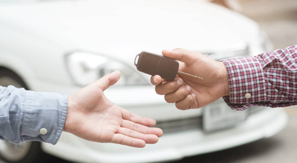 Man's arm wearing a plaid shirt handing a car key to another man's hand in front of a white car